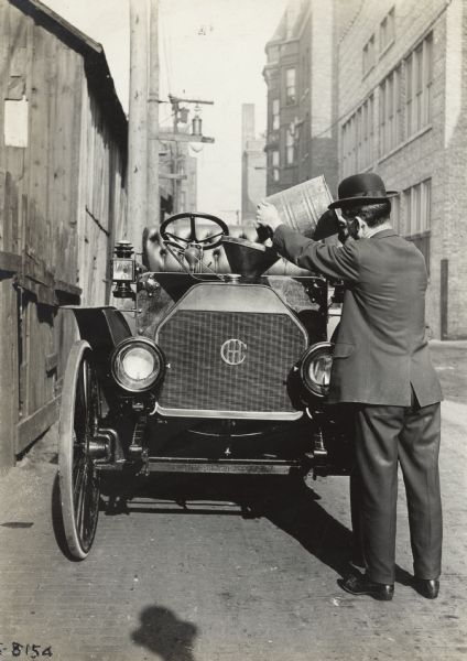 Well-dressed man standing in urban alley fueling an International Model M motor truck through a funnel.