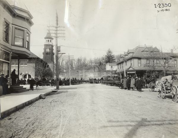 View down street of large group of men in small town with over 15 manure spreaders on display. On the left near an intersection is a firehouse with a tower, and on the right is the "Morton House."