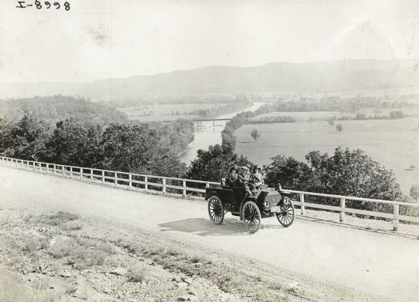 Slightly elevated view of a man driving three women in an International Model M truck on a rural road with a guard rail. In the background is a valley with a river and a bridge.