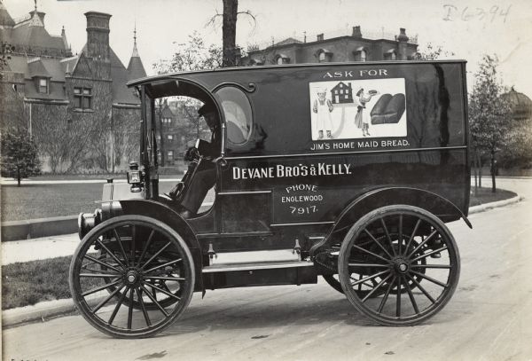 Man in driver's seat of an International Model M truck operated by  Devane Bros. & Kelly. Advertising on the side of the truck promotes "Jim's Home Maid Bread." In the background are large brick buildings surrounded by a lawn.