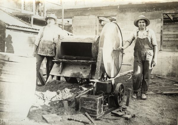 Two men posing at a construction site with an International stationary engine and concrete mixer. One of the men is wearing a work apron from Botsford Lumber, a lumber company operated out of Minnesota.