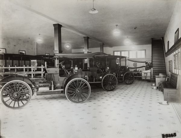 International trucks on display on a dealership showroom floor. There are three spittoons on the floor near benches along the wall. A stairway leads to a second floor. A Titan tractor is partly visible in the background.
