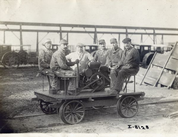 Several railroad workers sitting on a handcar with a Mogul engine mounted to it. Another man is standing just behind the group.