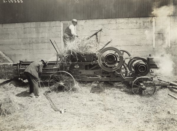 Men with Motor Hay Press Two men use a hay press to bale hay near a farm building. One man loads the press with hay while the other binds hay with twine. The press is powered by a small engine, probably an International Type M, mounted at one end.