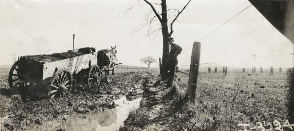 Panoramic view of man standing against fence near a horse-drawn wagon heavily loaded with dirt, with the wheels sunk deep in water-logged mud.