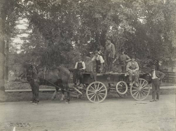 Group of men on and near a horse-drawn wagon on the side of a road. The wagon is loaded with tools and hardware. Four of the men wear tool belts.