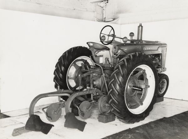 Farmall M tractor with an H&M 195 direct-connected Farmall plow on display in front of a white backdrop. The equipment may be experimental.