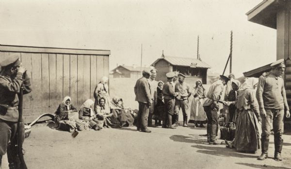 Group of men and women waiting outside small buildings, probably near a train station on the Trans-Siberian Railway. The stop may be somewhere between Novo-Nikaievsk and Irkutsk, Russia. Several women are seated on the ground with baskets, while a soldier in the foreground stands with a rifle. The original caption reads: "group of peasant women food vendors."