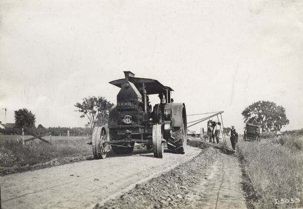 Large Mogul tractor pulling a grader along a dirt road. Two men ride in the tractor, while three others supervise from behind the tractor.
