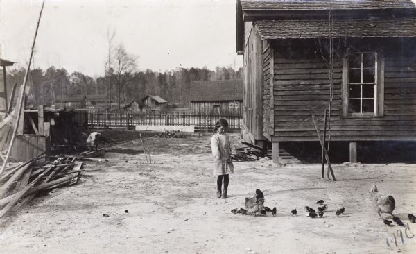 A girl wearing a dress and boots is standing outdoors in a yard near a house to feed chickens and chicks. In the background on the left a child is playing near stacks of lumber.