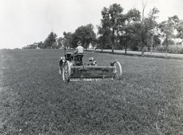 View from rear of a man using a Farmall tractor and a McCormick-Deering lime sower to fertilize an alfalfa field at International Harvester's Hinsdale experimental farm. There is a tree-lined road on the right.