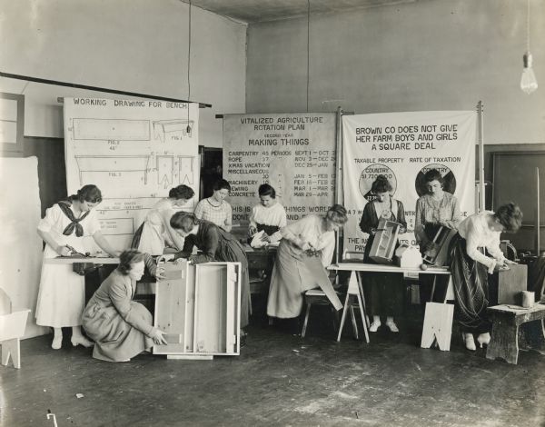 Group of women, probably teachers, using tools to assemble various wooden products including nail boxes, benches and cabinetry(?). Three large posters are hanging behind the women; one of them is titled: "Working Drawing For Bench". The teachers are likely learning projects for classroom use.