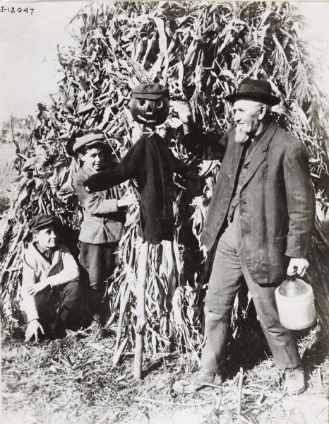 Older man and two young boys standing near a scarecrow made from a sweater, hat, and pumpkin erected near a shock of corn. The man is holding a jug.