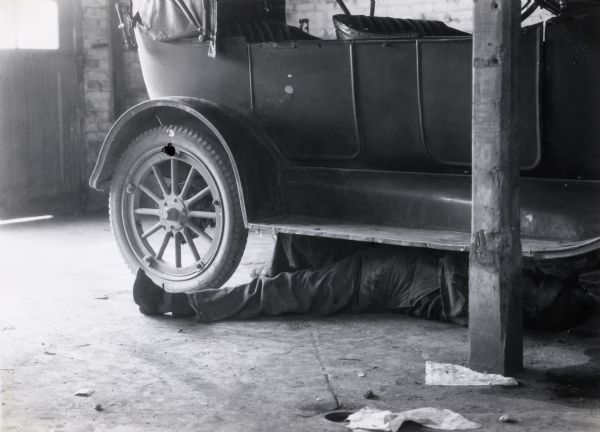 A man lies on the floor of a garage to repair an automobile at International Harvester's Hinsdale experimental farm. The photograph was taken for International Harvester's Agricultural Extension Department to warn of the dangers of working in a closed garage with an automobile engine running.