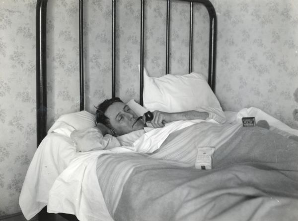A man lies in bed while smoking a pipe. A pouch of tobacco labeled: "Smoking Mixture" and a box of Red Top matches rest nearby on the blanket.
