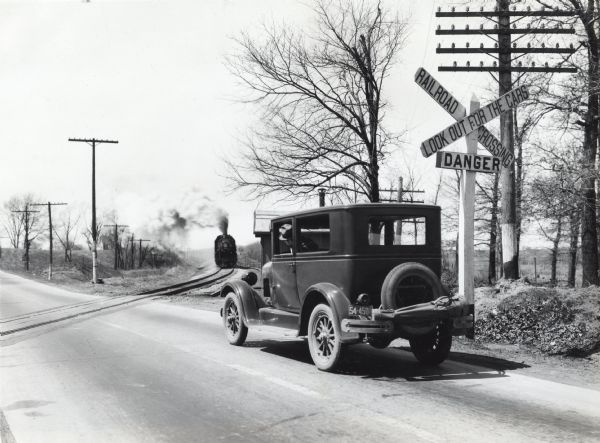 A man stops his automobile at a railroad crossing while waiting for the train to cross a paved road. The traffic sign to the right of the car reads: "Railroad Crossing," "Look Out For the Cars," and "Danger."