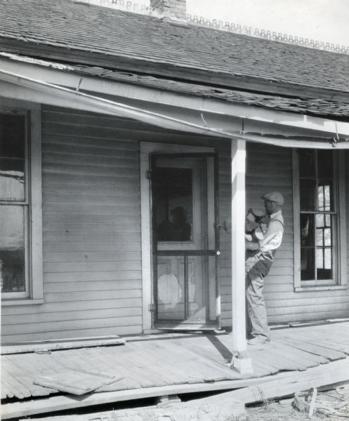 A man holding a bundle of logs is opening the screen door on a farmhouse with a deteriorating front porch.