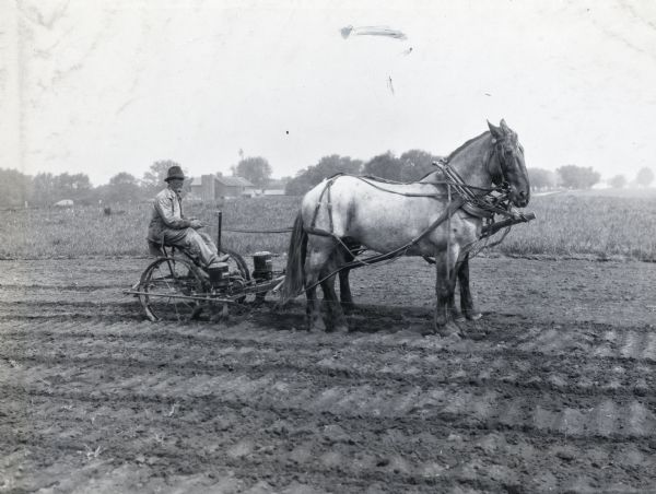 A man is smoking a pipe while operating a McCormick-Deering a horse-drawn corn planter in a farm field. A silo, barn, and other farm buildings are in the background.