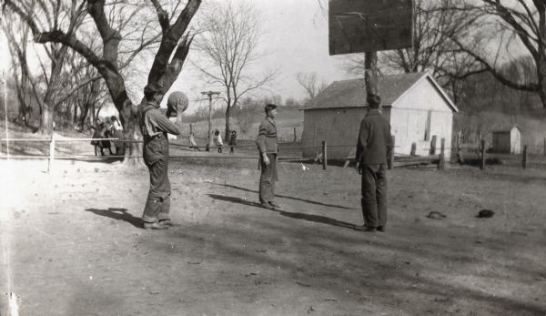 Three young men playing basketball on an outdoor court. In the background behind a fence and tree are a group of girls, and in the center background younger girls are playing on playground equipment near a building, possibly a school.