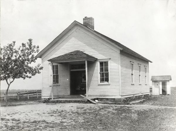 Exterior view of the front of a one-room schoolhouse. There is a Red Cross poster hanging in the school entryway, and a smaller outbuilding behind the schoolhouse.