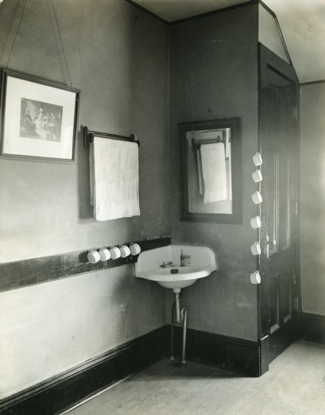 View of a sink installed in a corner in a washroom at School #9. A mirror, towel rack, framed picture, and a row of mugs hang from walls surrounding the sink.