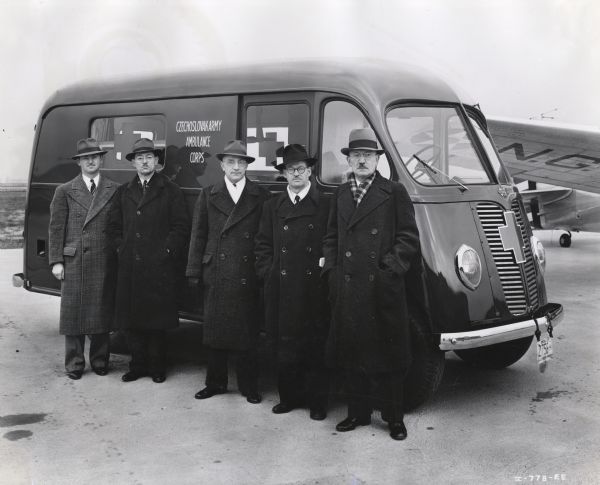 A group of men from the Chicago Czechoslovak Relief Committee are standing against an International D-15-M (Metro) truck sold to Czechoslovakian Relief for service with the Czechoslovakian Army in England. An airplane is in the background on the right.
