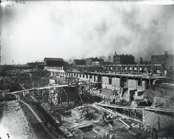 Elevated view of a construction site surrounded by buildings in what appears to be an industrial area. A sign on the building on the left side of reads: "Woolf Furnishes Your House in One Day's Notice for Cash."