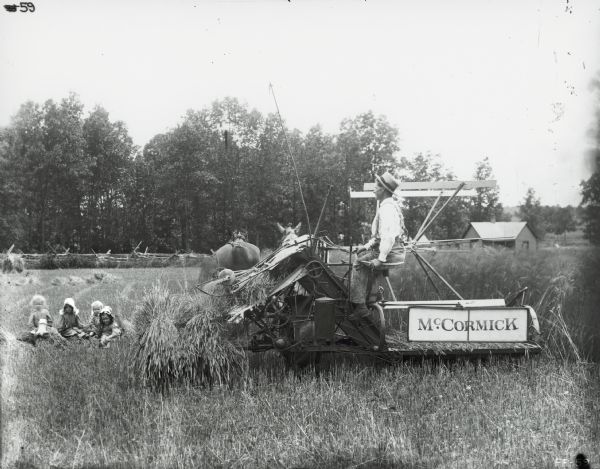 Four small children are sitting on shocks of wheat in a field as a man is operating a McCormick grain binder nearby. Farm buildings and a fence are in the background.