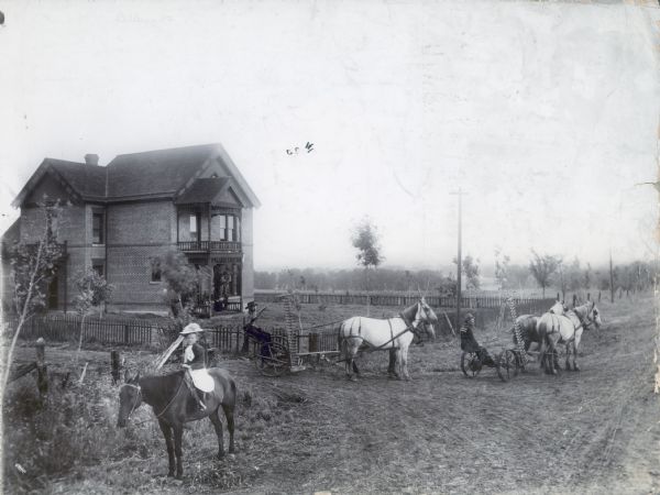 Two men are leading horses in pulling McCormick Big 4 mowers near a brick farmhouse. A girl wearing a dress and hat is sitting on a horse in the foreground.