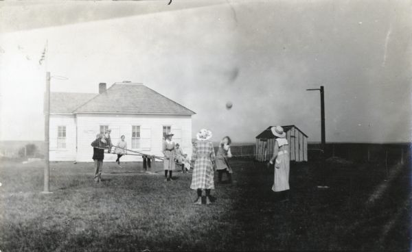 A group of children playing volleyball during recess outside of a school building. Additional children are playing on a see-saw behind them.