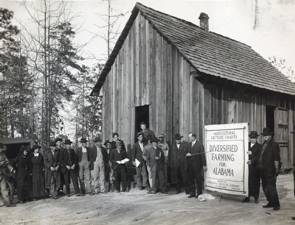A group of men and women standing outside the New Home School to listen as Professor Clark is speaking on the subject of diversified farming. He uses a pointer to direct attention toward a visual aid that reads: "Agricultural Lecture Charts. Diversified Farming for Alabama. Prepared by International Harvester Company of New Jersey. Chicago."
