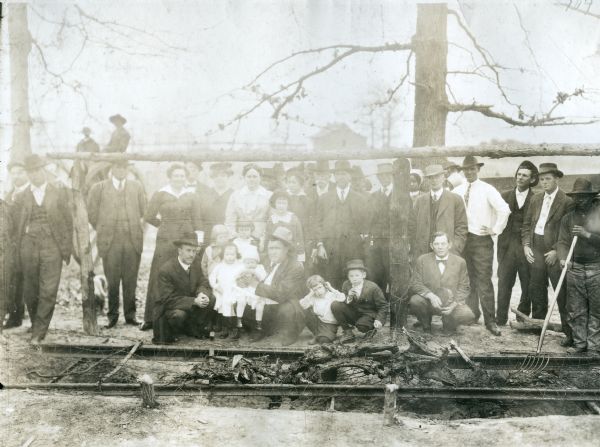 A group of men, women, and children gathered around an outdoor barbecue pit as meat roasts. The barbecue took place at Louis Frank Sessions' farm during a meeting on diversified farming.