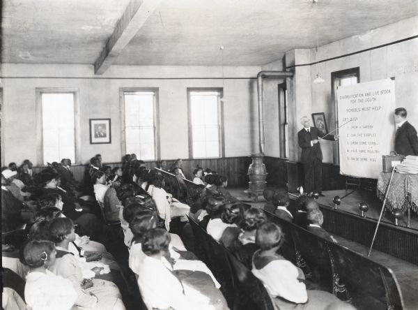 Professor C.W. Farr standing on an elevated platform at the front of a classroom at the Burrell School while presenting on the subject of agricultural diversification. He is using a pointer to direct his class' attention to a visual aid as Principal White is standing beside him. Audience members are sitting in rows of chairs throughout the classroom.