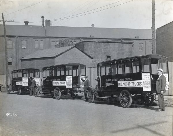 Three men stand next to International Model F (or 31) buses parked along the side of a street. Two men are sitting in the driver's sseat of two of the buses. The text on the buses reads: "DeGraff Village Union Schools" and the attached signs read: "I.H.C. Motor Trucks. Firestone Tires Akron O. on this, as on thousands of other trucks Throughout the World."