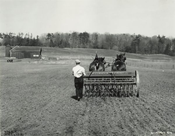 McCormick-Deering Grain Drill in Field Man walks behind a horse-drawn McCormick-Deering grain drill in a field. Decals are on the grain drill. Barns and other farm buildings are in the background.