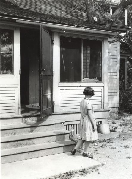 A girl wearing a dress and "Mary-Jane" shoes walking across the cement stair landing of the farmhouse entrance at International Harvester's demonstration farm. The screen door to the enclosed porch of the house is standing open.