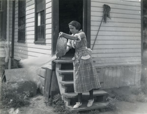 Woman dumping wash water from a metal wash basin on the back step of her farmhouse.