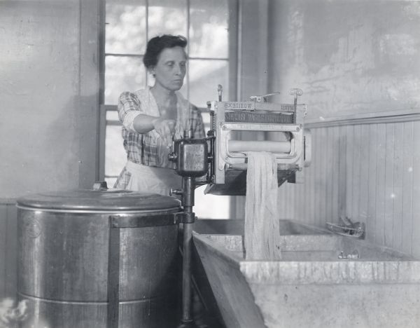 A woman wearing a dress and apron is using an "Easy Model H" wringer washing machine made by the Syracuse Washer Corporation.