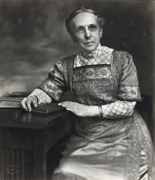 Three-quarter length portrait of Miss Ella Flagg Young seated at a desk. She is wearing a patterned dress and has her hands on a book. Ella Flagg Young was the first woman in America to head a large city school system.