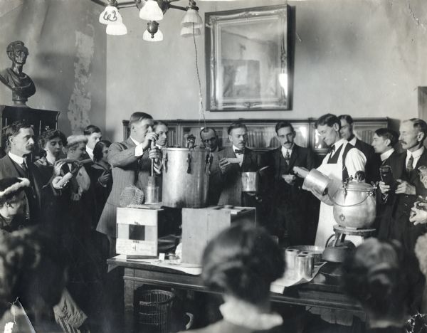 A group of men and women surround O.H. Benson as he gives a canning demonstration in the office of County Superintendent Tobin. Canning equipment stands on a table and bookshelves are in the background behind the spectators.