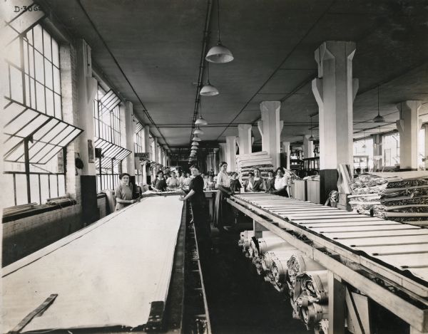 Female factory workers stand around long tables in the canvas department at International Harvester's Deering Works Canvas Department. Canvas is piled in rolls beneath the tables and folded in large stacks. The canvas was likely used for grain binders, harvester-threshers (combines) and push machines.