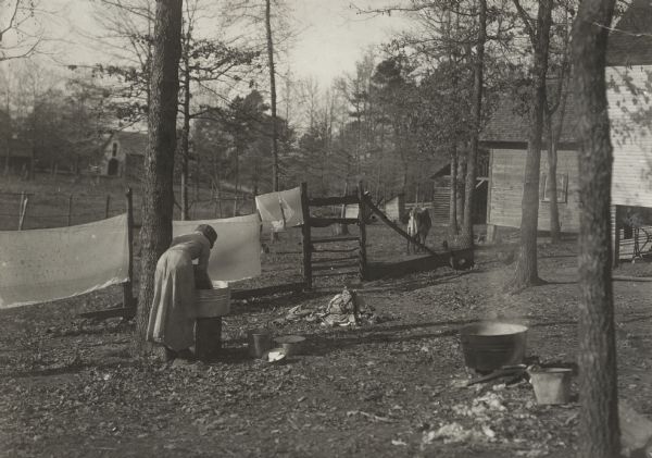 View across wooded yard towards a woman leaning over a washtub using a washboard and to wash sheets. Sheets are drying on a clothesline and a washtub of boiling water is in the foreground.