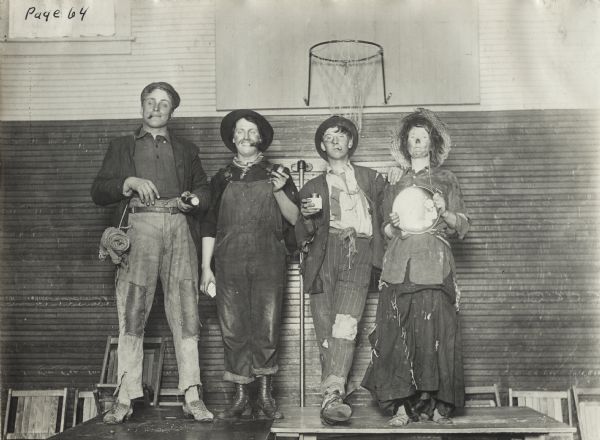 Young men and women standing on a platform in a school gymnasium dressed as hobos for a "Hobo" party at Woldale Rural School. A basketball hoop is in the background.