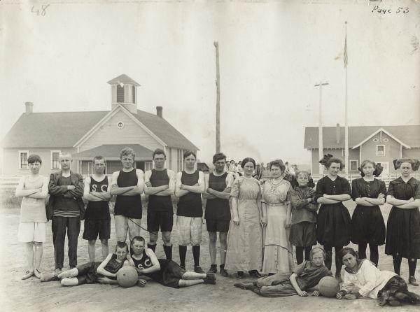 Group portrait of boys and girls dressed in basketball uniforms with teachers outside a gymnasium (at right) at the Woldale Rural School.