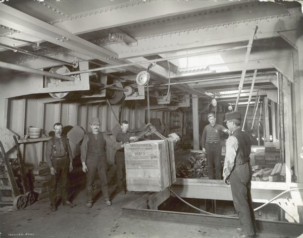 Factory workers are loading a wooden crate marked "McCormick Folding Daisy Reaper" using a pulley system in the docks of the McCormick Works.