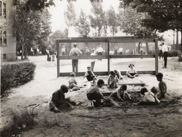 Children playing games behind a fence. In the foreground other children are playing in a sandbox on a school playground next to several buildings.