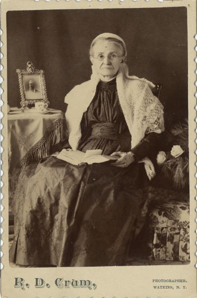 Carte-de-visite of Amanda Joanna McCormick Adams seated in parlor-like setting on Christmas. Mrs. Adams is seated before a framed photograph of a man on the table next to her, possibly husband Hugh Adams. She sits in a chair with an opened book in her lap.