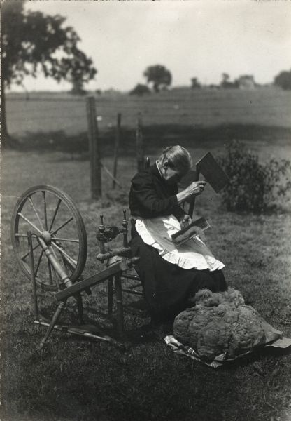 A woman sitting outdoors next to a spinning wheel while carding wool.