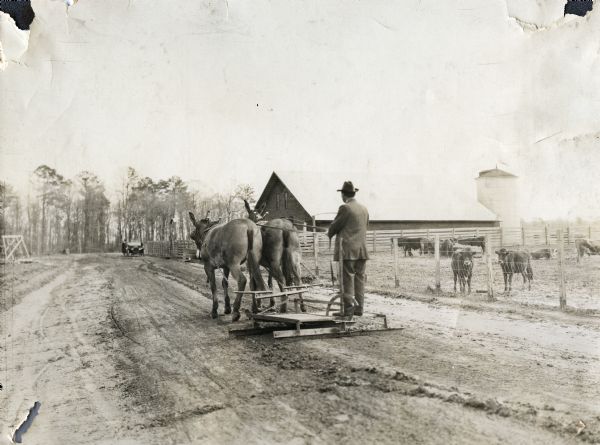 A man wearing a suit and hat is standing on a wooden grader while driving two mules along a dirt road. Beside the road behind him are cows in a fenced-in field near a barn and silo.