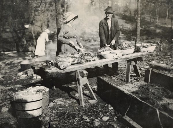 Mr. and Mrs. T.J. Crumley cutting slabs of meat from a hog placed on a wooden table outdoors among trees. Another woman is in the background near some steaming pots, and a bushel basket and pail are standing near the table.